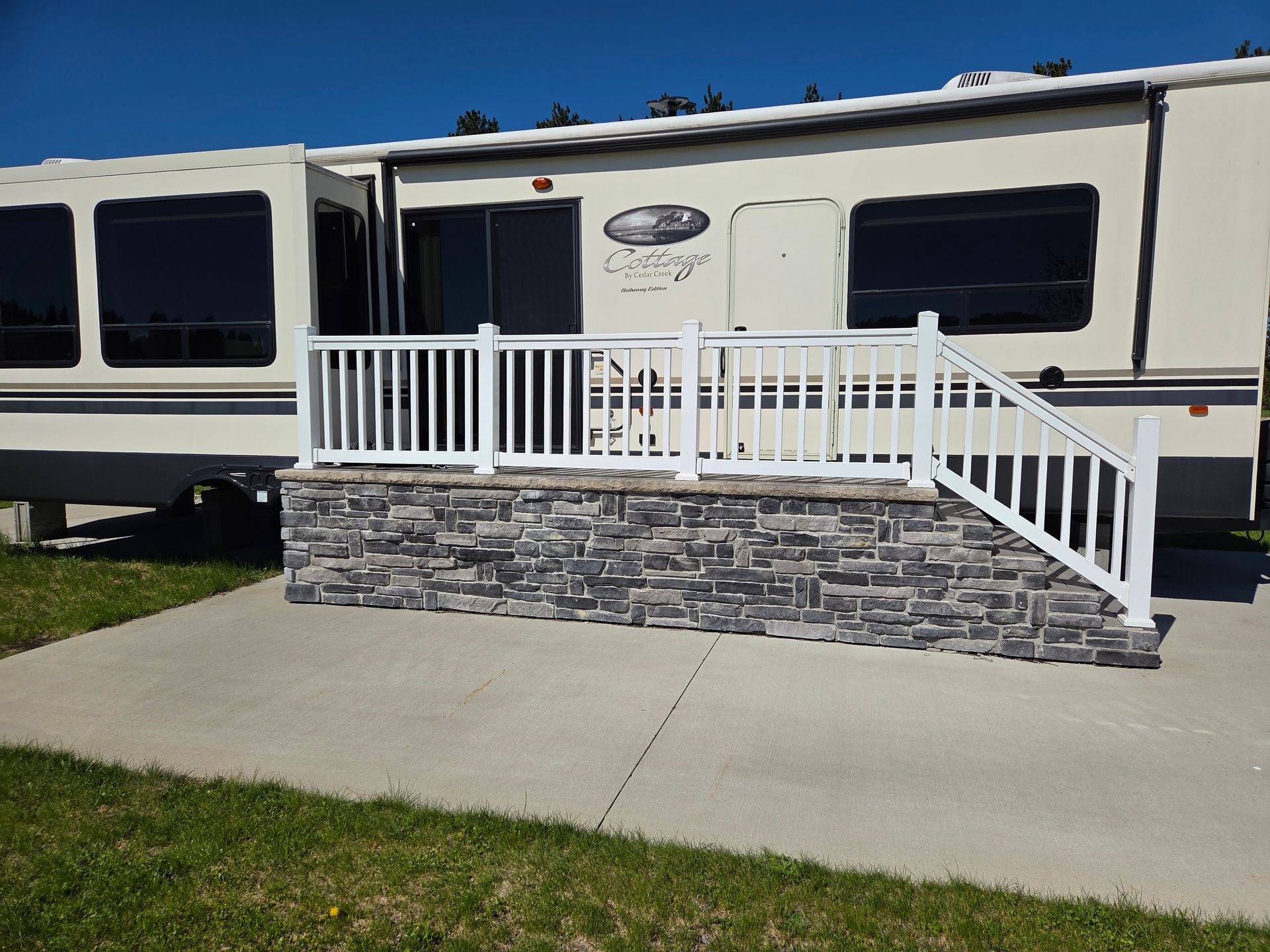 White RV with a small stone-front porch, white railing, and concrete steps on a grassy lot.