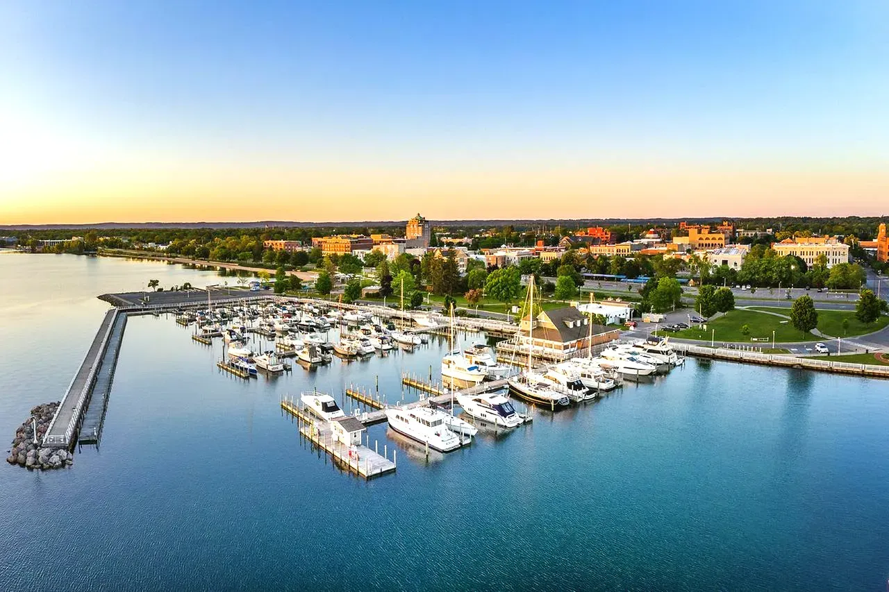 Boats docked in a harbor, city buildings in the background, blue water, and a clear sky.