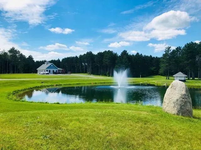 Lush green landscape with pond and fountain, house and trees under a blue sky.