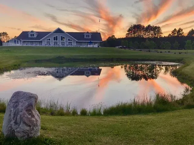 Pond reflecting a building under a colorful sunset sky. A large rock sits in the foreground, and trees line the horizon.