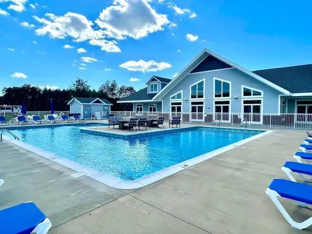 Swimming pool with blue water and surrounding lounge chairs, beside a light blue building under a bright, cloudy sky.