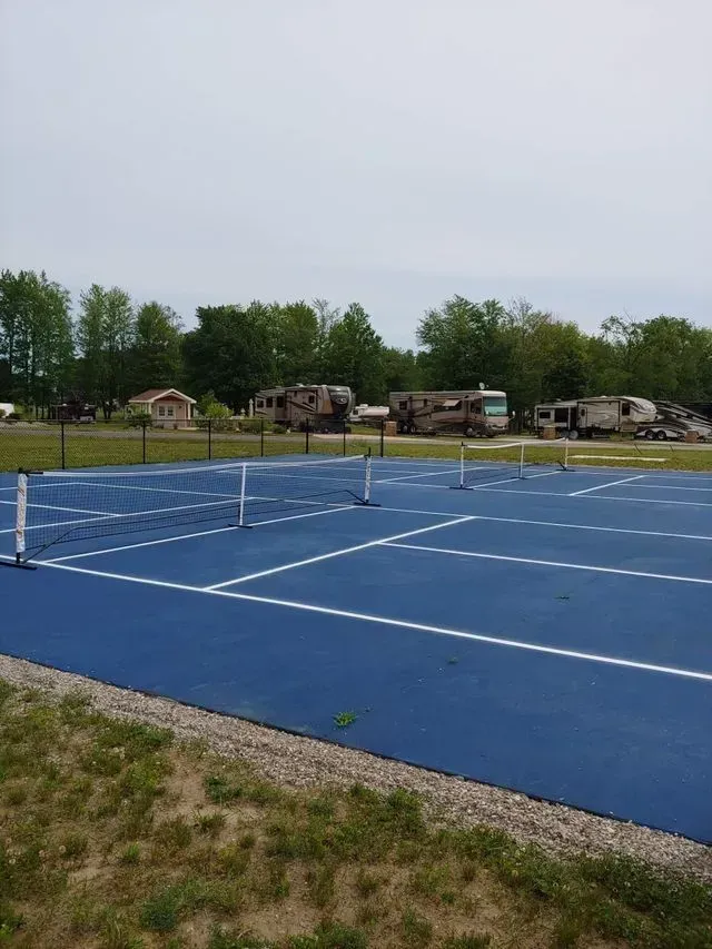 Blue tennis court with RVs in the background, set in a grassy area with trees, overcast sky.