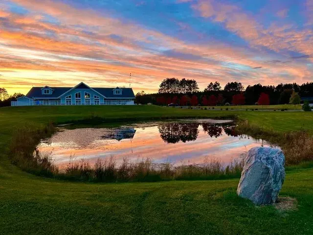 Pond reflects a colorful sunset and a blue building on green grass, with a large rock in the foreground.