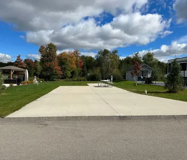 A concrete RV pad in a campground with grass, trees, and cloudy blue sky.