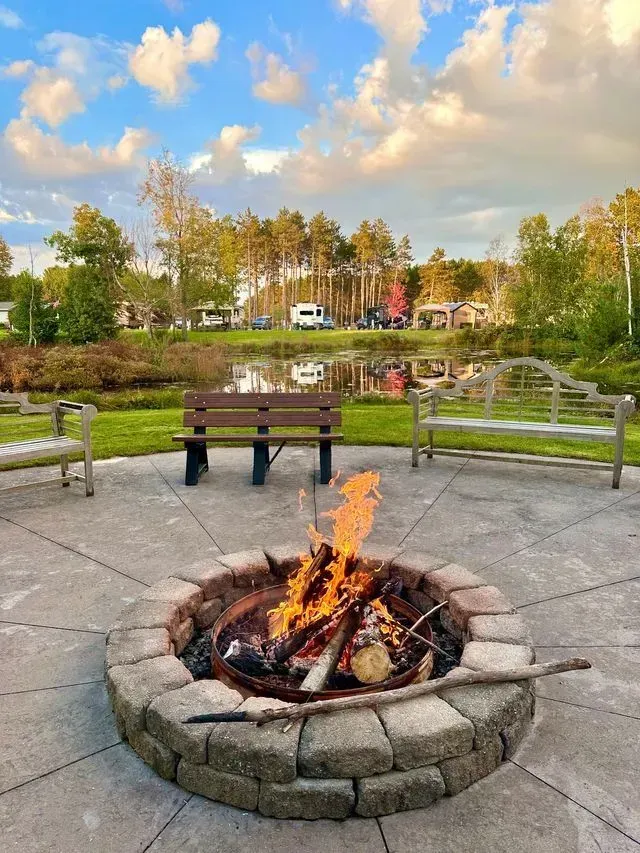 Fire pit on a circular patio, benches around. Campground with lake and trees in the background under a cloudy sky.