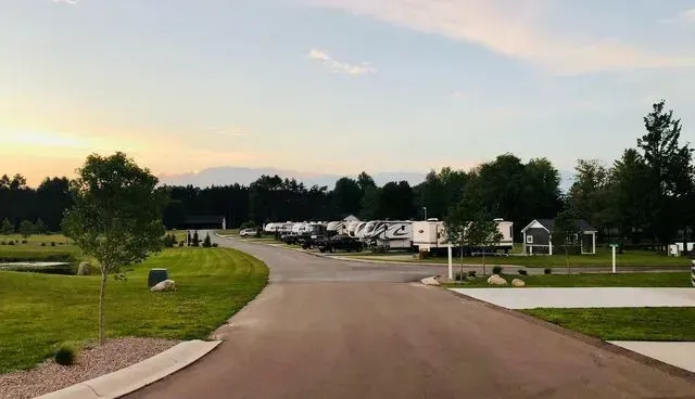 Paved road through a campground with RVs and grassy areas, trees, and a light blue and orange sky.