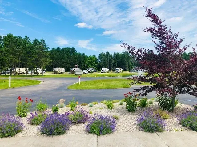Campsite with RVs, green grass, trees, and lavender bushes under a blue sky with clouds.