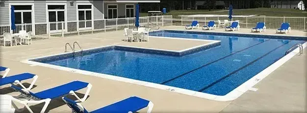 Swimming pool with blue water and surrounding concrete deck; lounge chairs and building in the background.