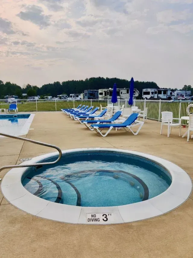 A hot tub and lounge chairs at a pool with a campground in the background. Blue umbrellas offer shade.