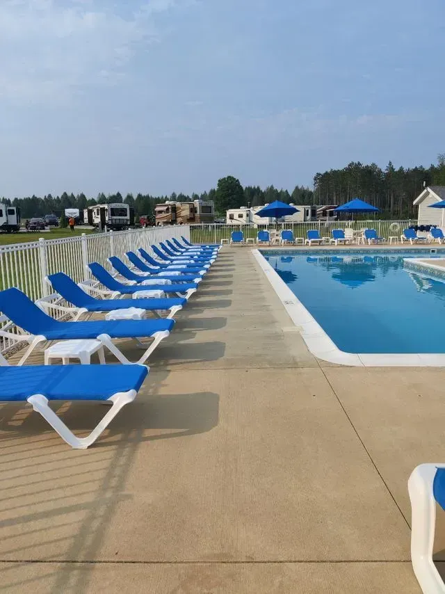 Row of blue lounge chairs beside a swimming pool; RVs and trees in the background.