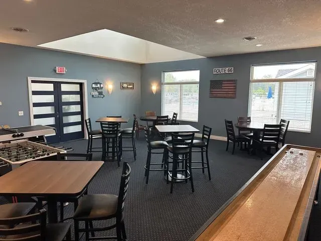 Game room with tables, chairs, foosball, and shuffleboard; blue walls, windows, and an American flag.