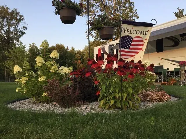 RV campsite with flowers, flag, and hanging plants; green grass, gravel border, dusk lighting.