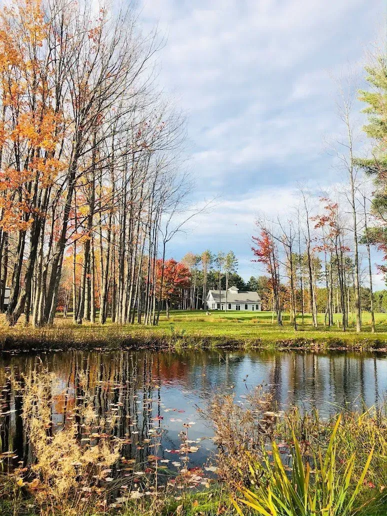 Pond reflecting fall trees with a white building in the background. Cloudy sky overhead.