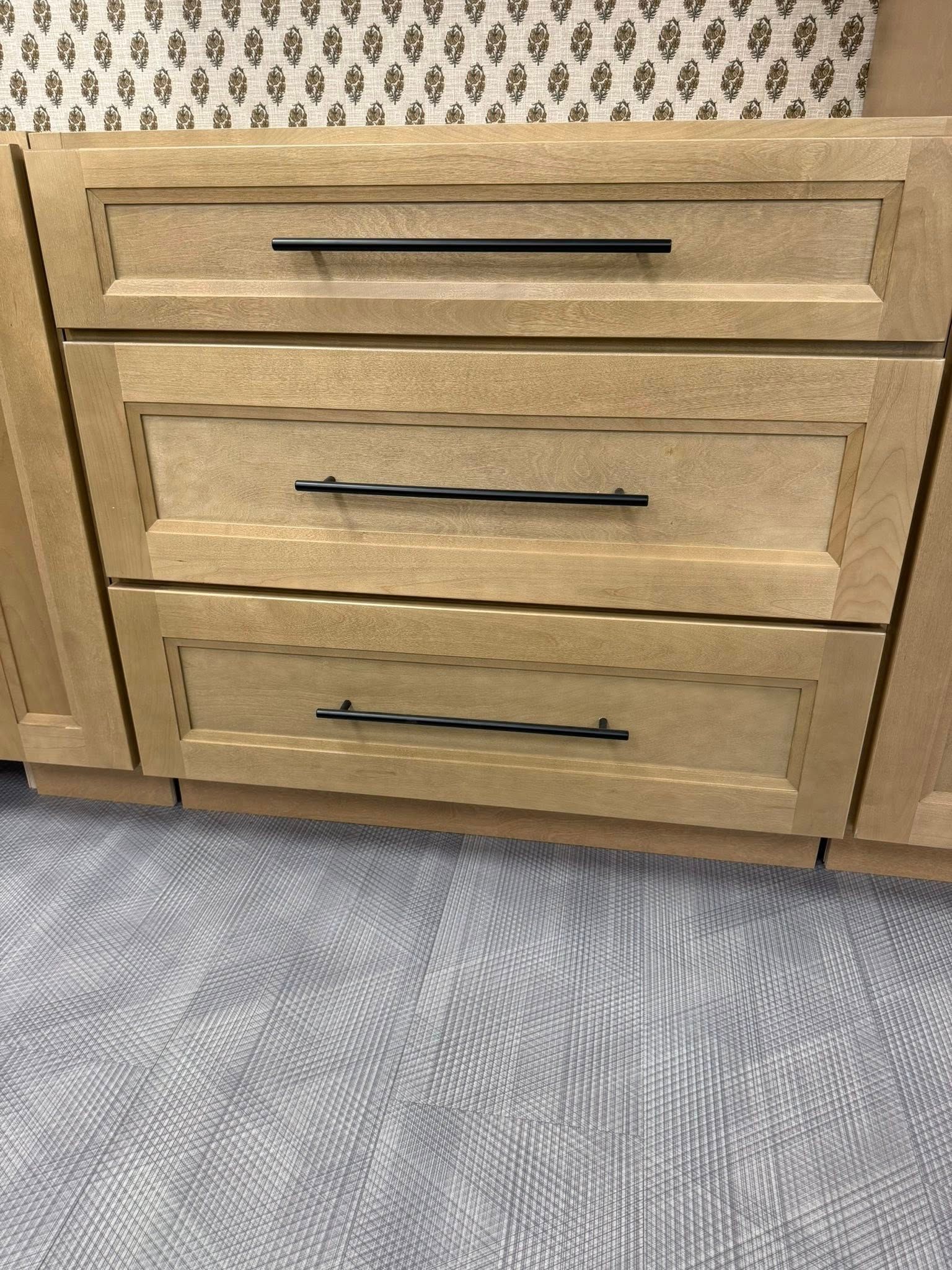 Three tan kitchen drawers with black handles against a patterned backsplash, sitting above a patterned gray carpet.