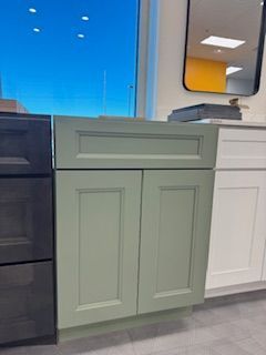 Sage green kitchen cabinet with two doors and a top drawer, shown in a showroom setting.