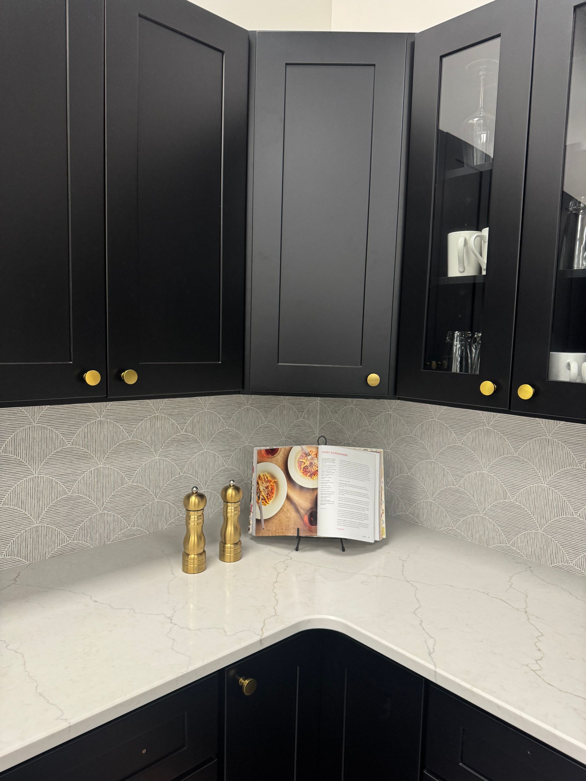 Black kitchen cabinets with gold knobs, white countertops, and patterned backsplash. Cookbook and spice set displayed.