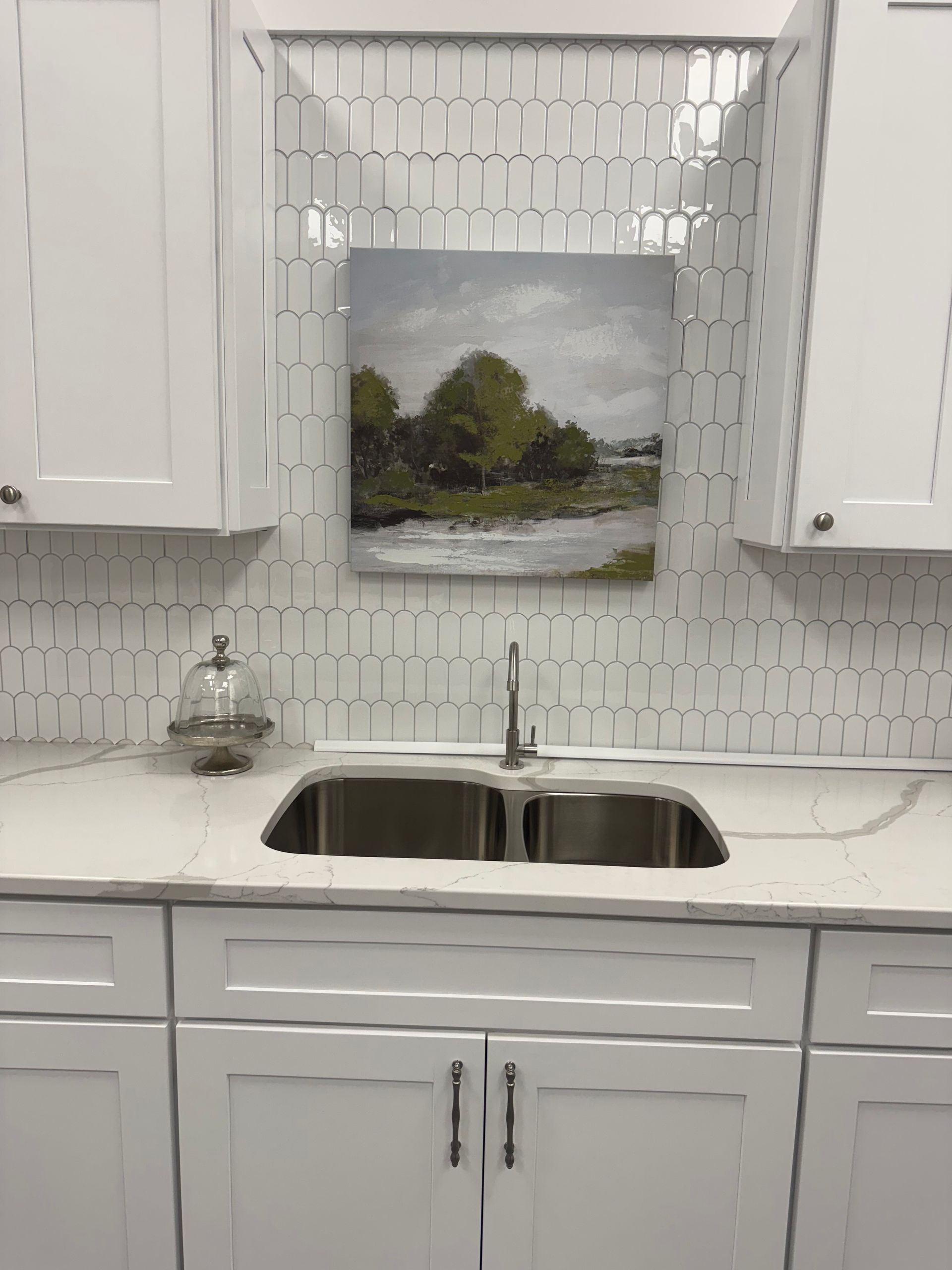White kitchen with a stainless steel sink, countertop, and cabinets. A painting hangs on the tiled backsplash.