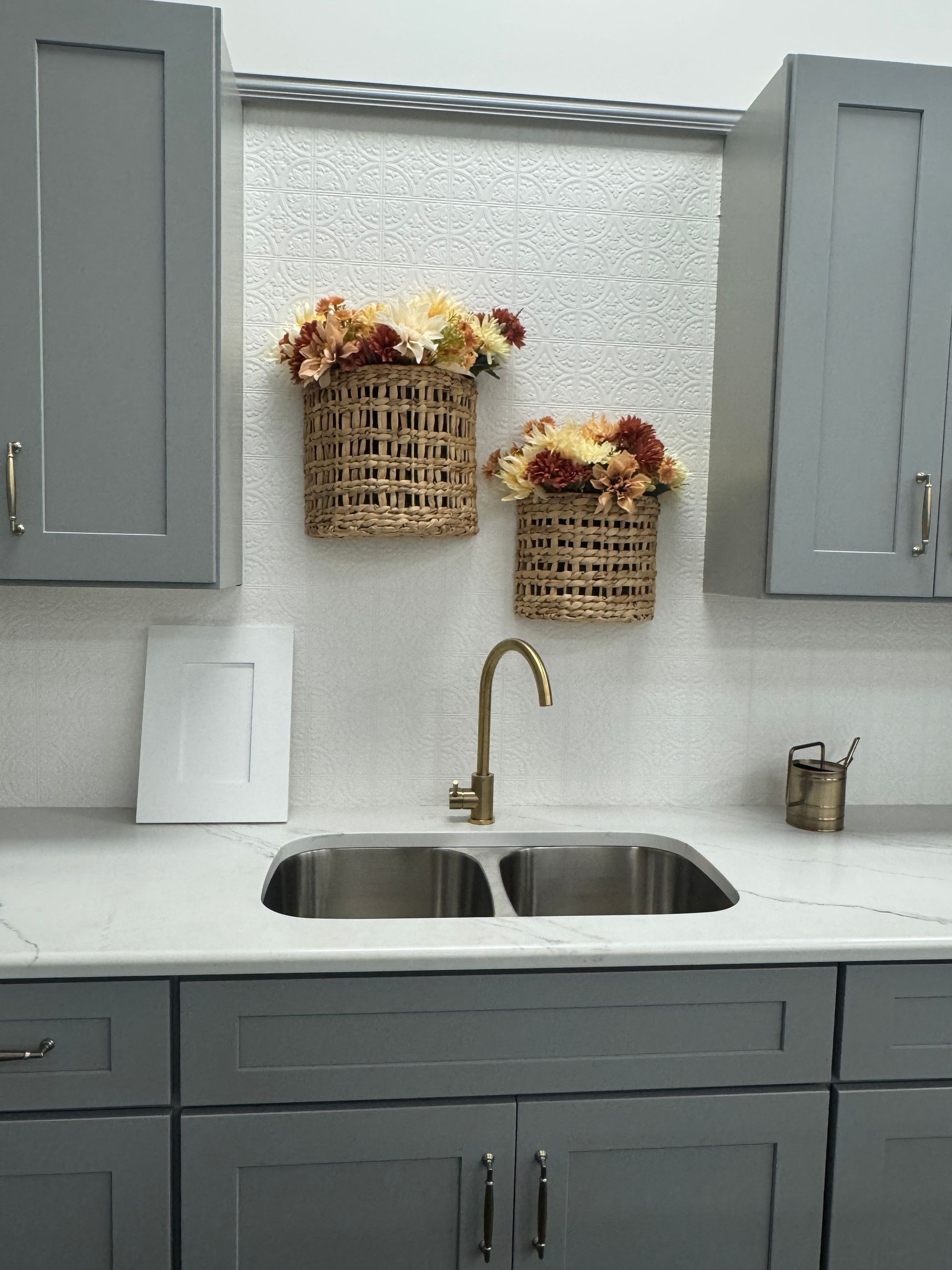 Gray kitchen with double sink, gold faucet, baskets of flowers, and white countertop.