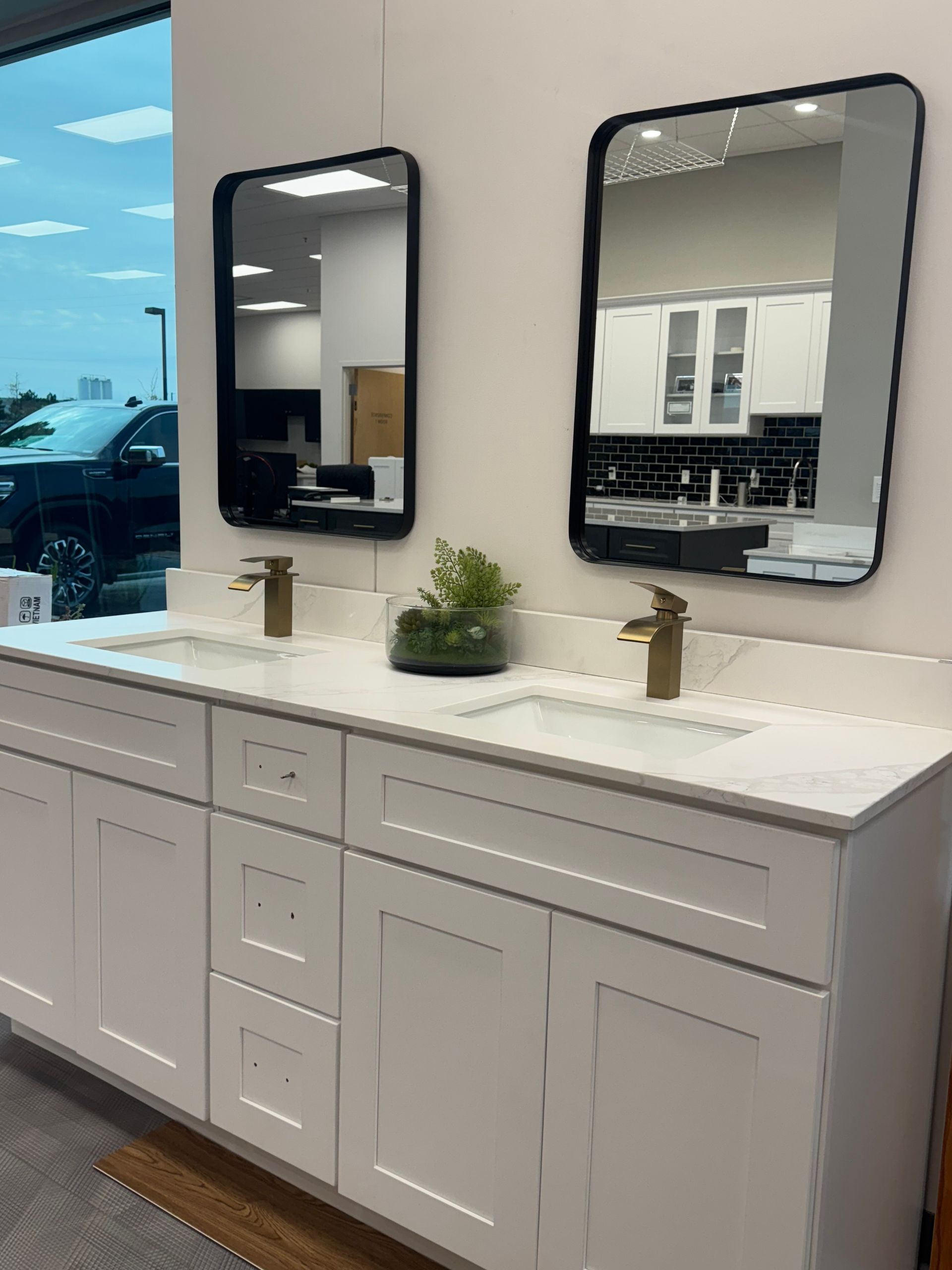 White bathroom vanity with two sinks, gold faucets, black mirrors, and a plant.