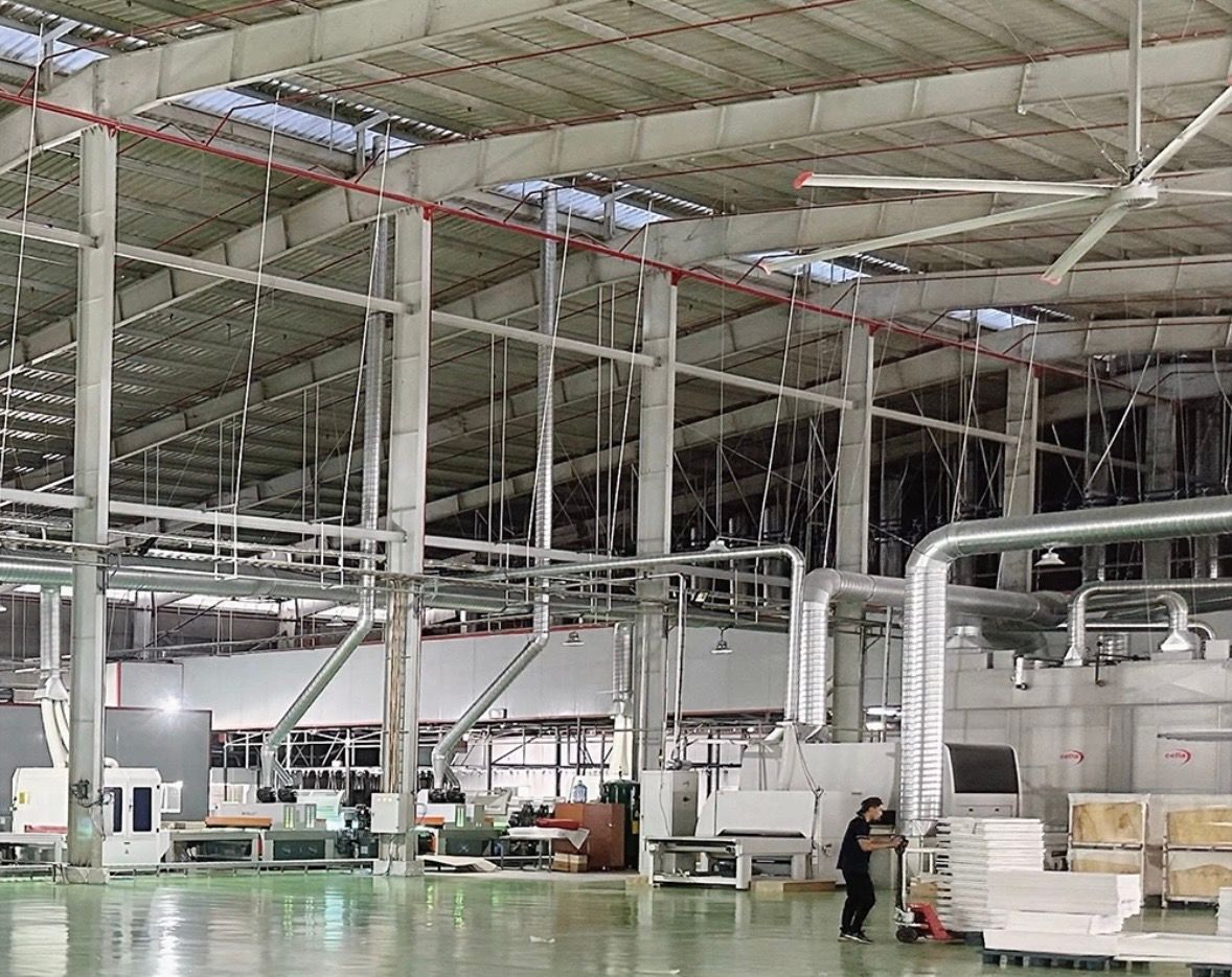 A spacious industrial warehouse with steel beams, overhead ducting, a large fan, and a worker operating machinery.