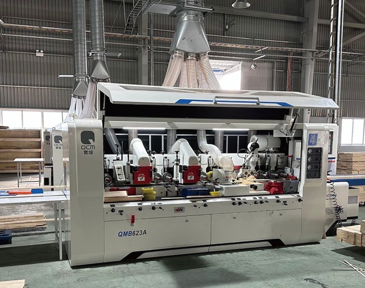 A white industrial woodworking machine with multiple cutter heads and overhead dust extraction ducts in a workshop.
