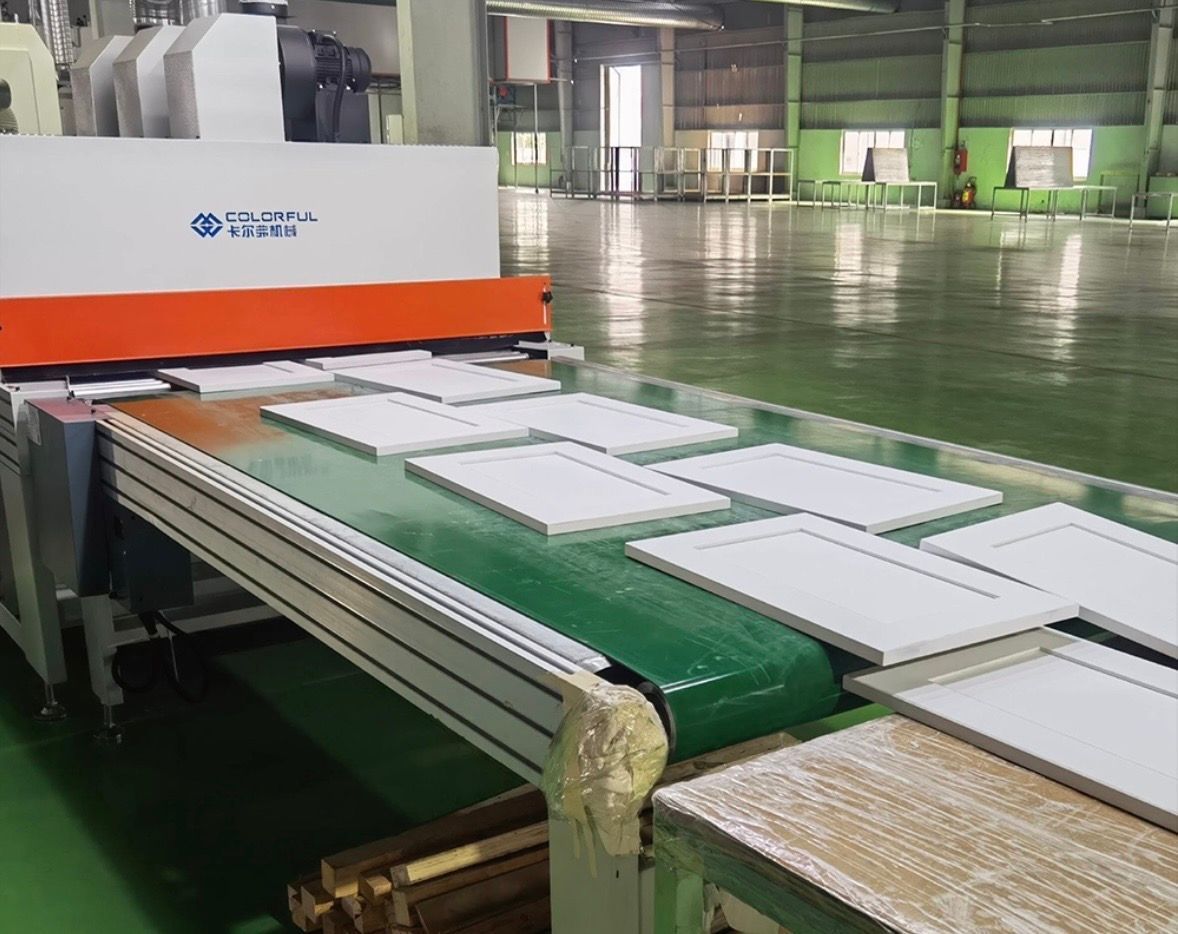White boards move along a green conveyor belt toward an industrial painting machine in a brightly lit factory.