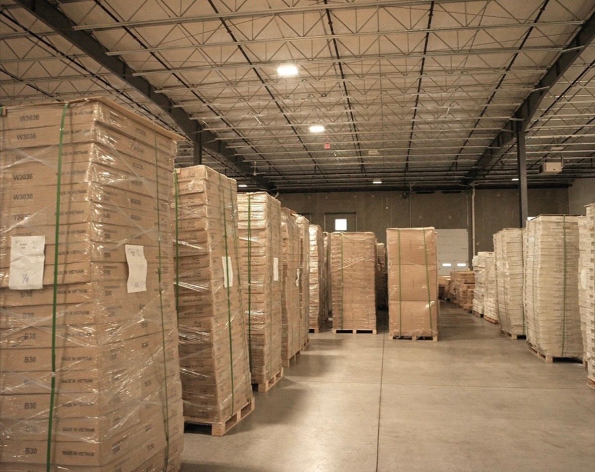 Rows of stacked cardboard boxes wrapped in plastic on wooden pallets inside a large, industrial warehouse.