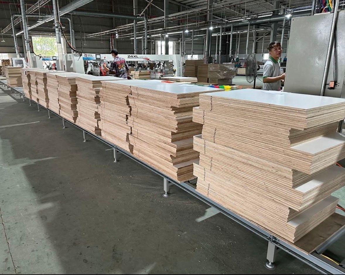 Stacked sheets of layered plywood with white finishes move along a metal conveyor belt in a large manufacturing factory.