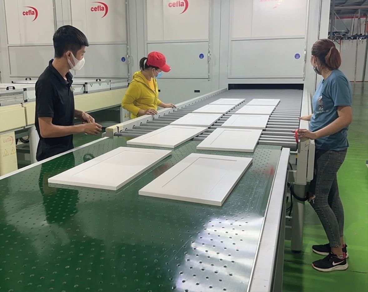 Three workers in a factory environment monitor flat white panels moving along a conveyor belt.