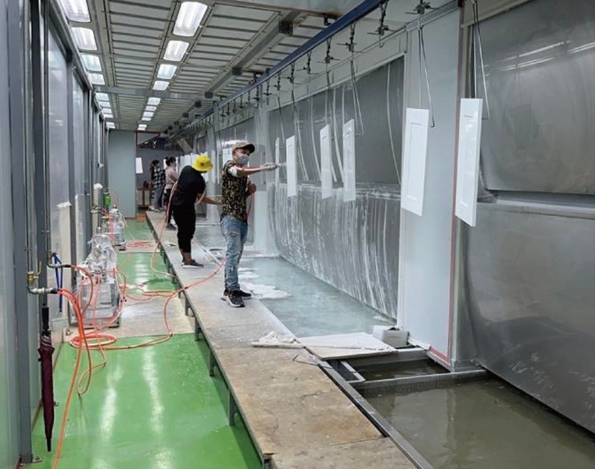 Workers in protective gear apply white, rectangular materials to a large, metallic wall surface in a bright workshop.