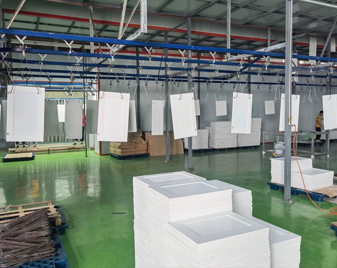 White panels hang from a conveyor system in a large warehouse with a green floor, with stacks of panels on the ground.