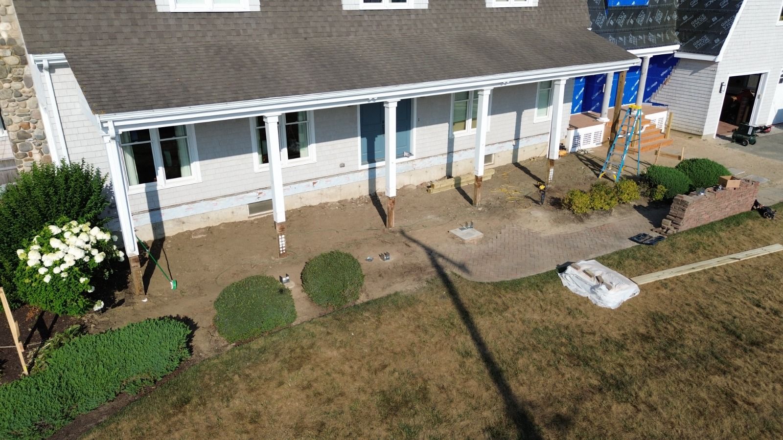 Gray house with white pillars, porch, and brown yard; bushes and a small garden.