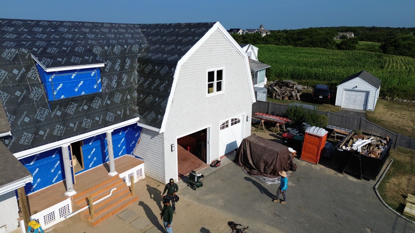 Construction site: Two-story house with gray siding, barn roof, and workers. Blue wrap covers some walls; sunny day.