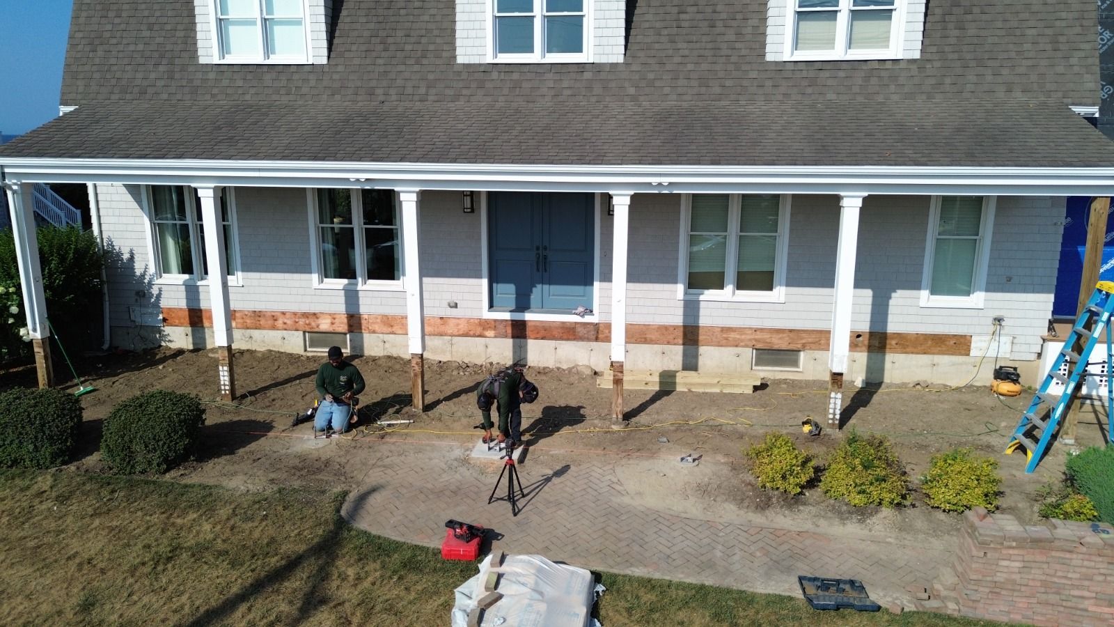 Workers replacing supports under a house's porch. Front of the house, light gray siding, blue door, gray roof.