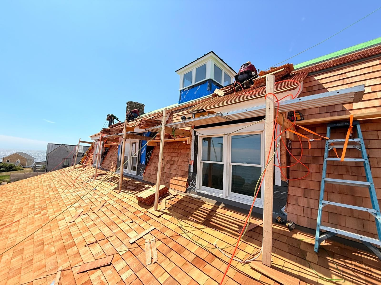 Roofers working on a wood shingle roof, blue sky, a dormer, and a ladder.