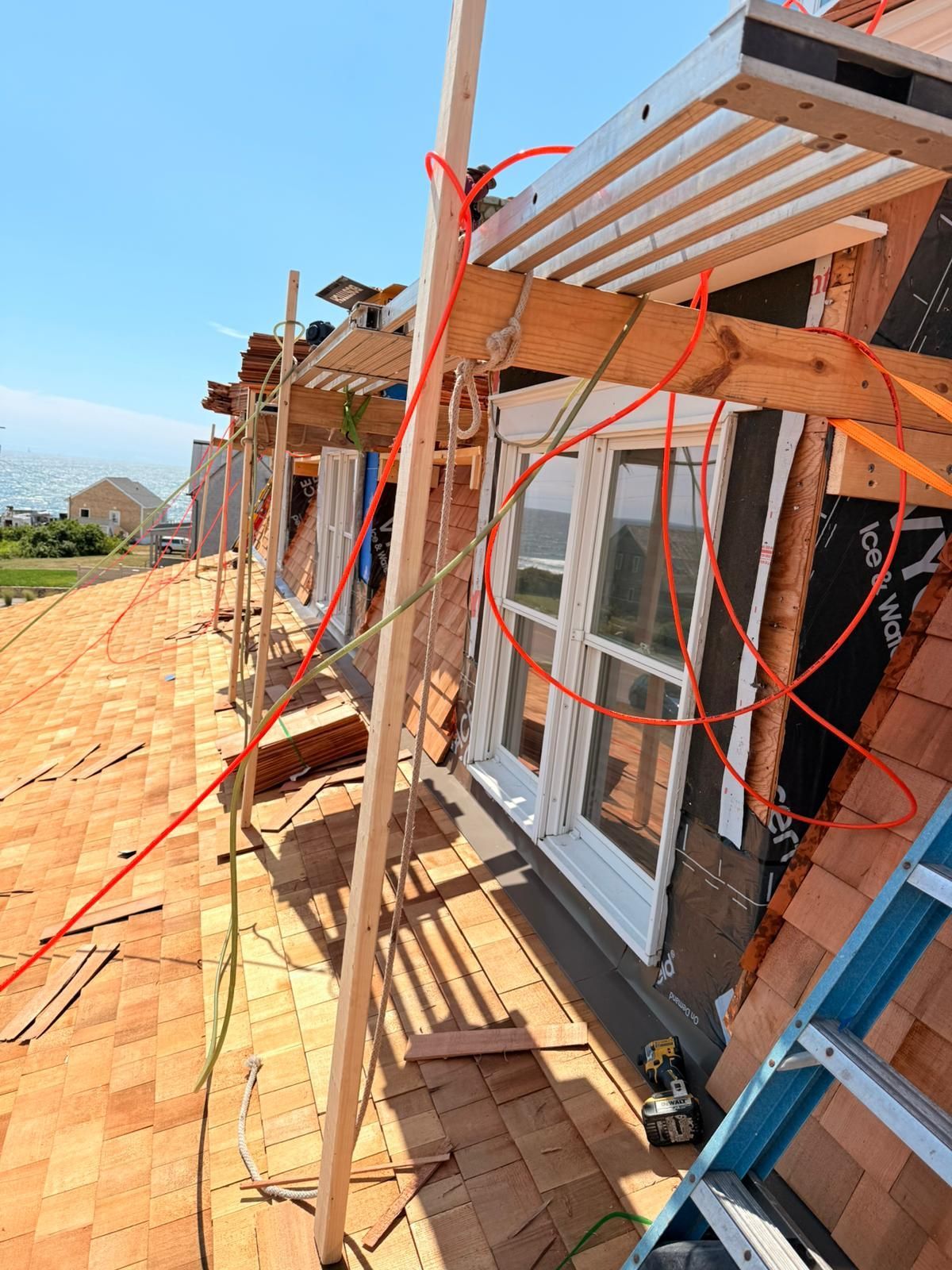 Construction site with workers installing roofing shingles. Scaffolding and electrical cords visible.