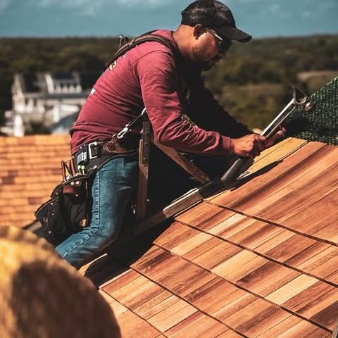 Roofer in burgundy shirt and hat hammers wood shingles on a house roof.