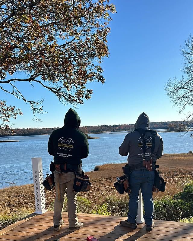Two people in hoodies, tool belts facing a body of water, under a tree.