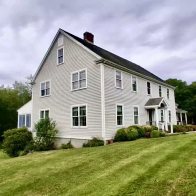 Two-story light grey house with white trim, black roof, and a grassy lawn under a cloudy sky.
