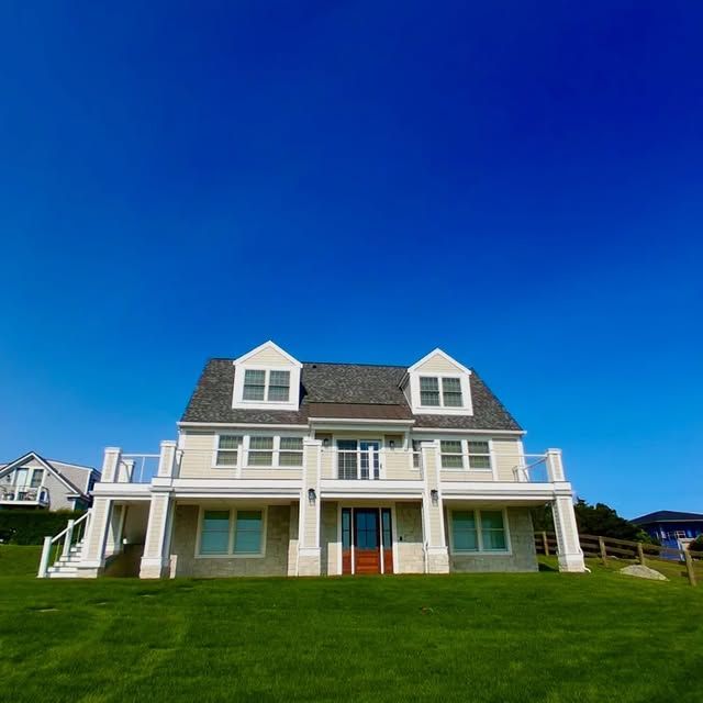 Two-story beige house with white accents, brown door, two dormers, and a balcony on a grassy hill under a clear blue sky.