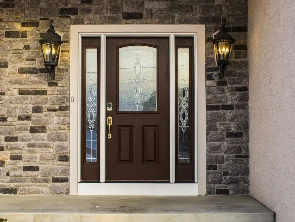Brown front door with glass panels, flanked by sidelights and lanterns, set in a stone wall exterior.