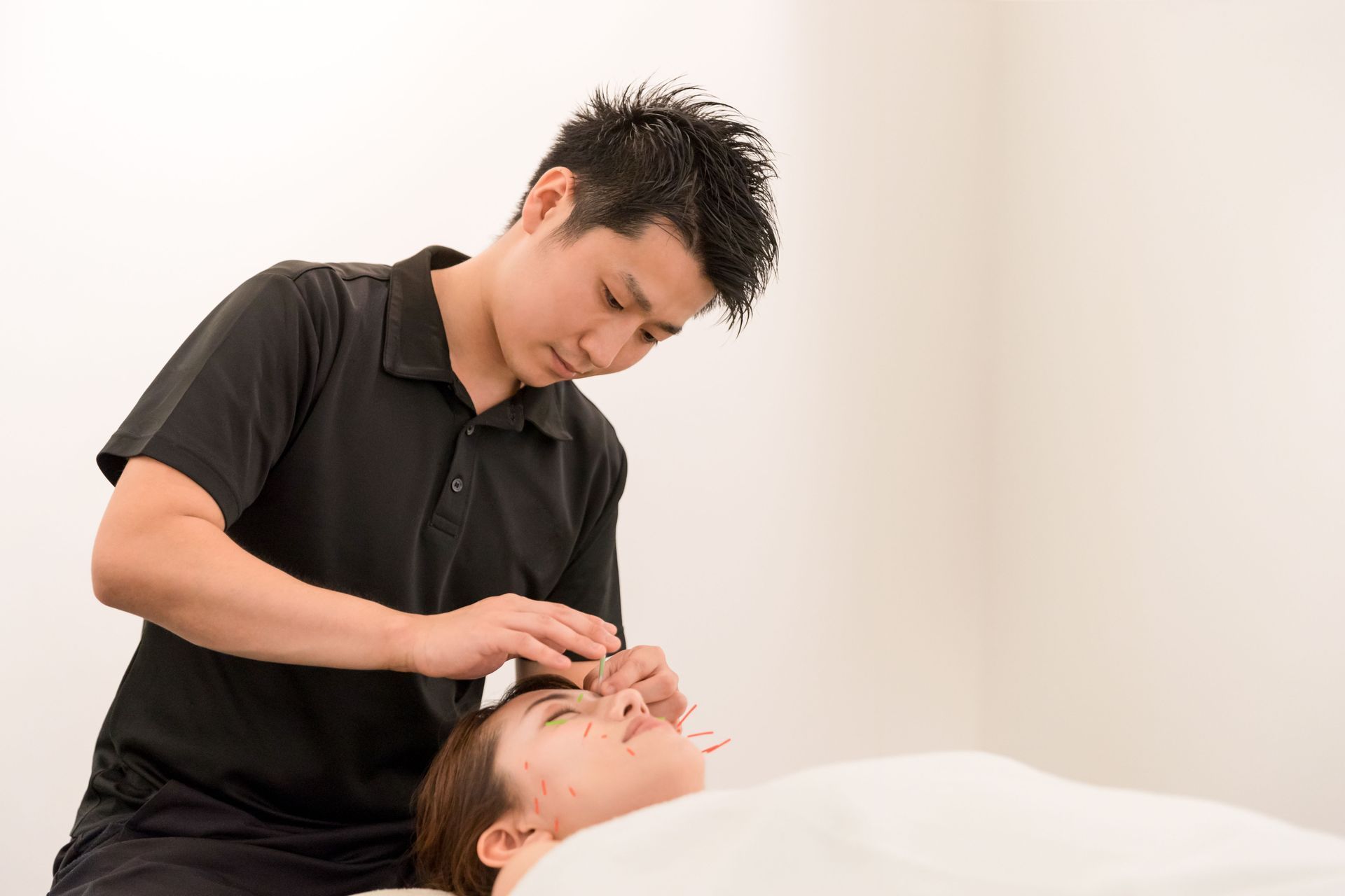 A therapist in a black shirt performs facial acupuncture on a person lying down in a bright, clinical space.
