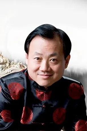 A man smiling while wearing a black jacket with a red circular pattern, set against a plain light background.