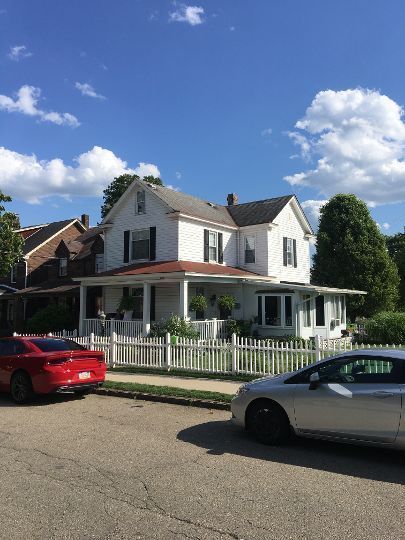 White two-story house with black shutters, red roof, and white picket fence on a sunny day. Cars parked in front.