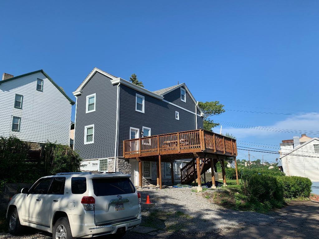 Two-story house with a wooden deck, dark gray siding, and a white SUV parked in front on a sunny day.