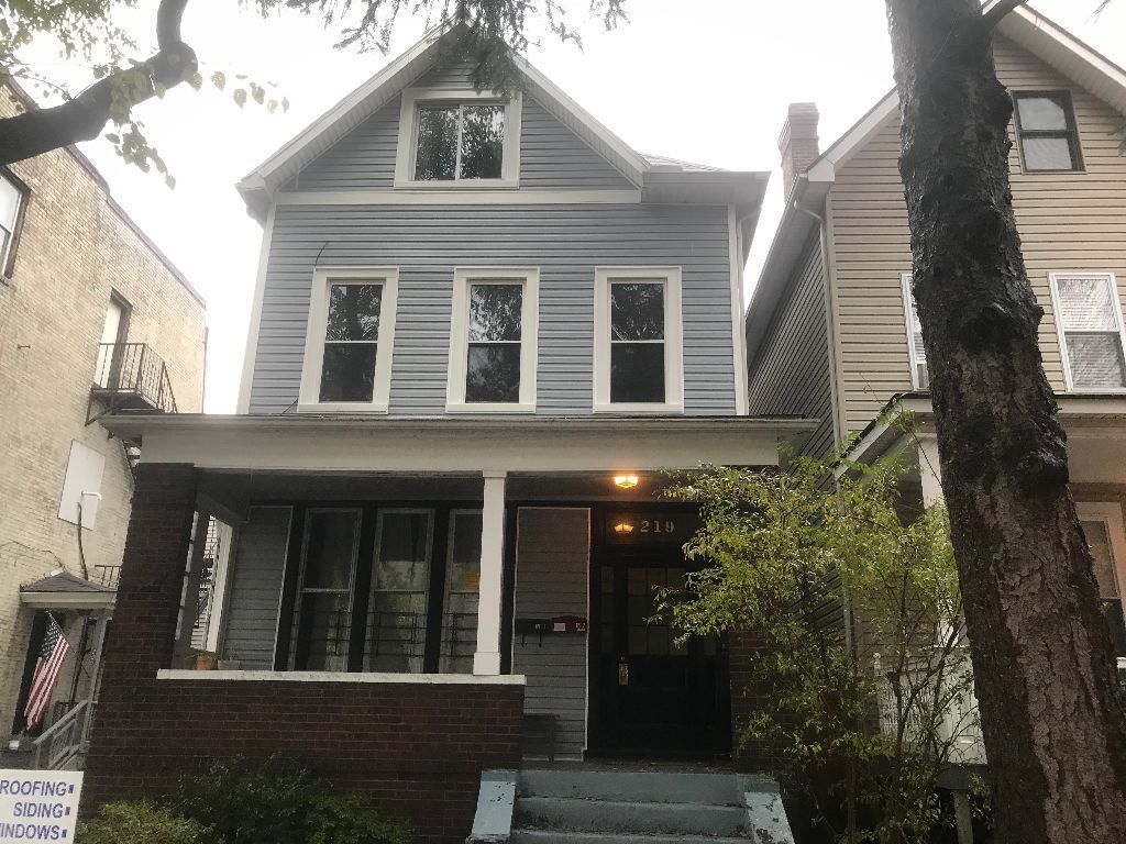 Three-story blue house with porch, windows, and steps. Trees frame the sides, a cloudy sky overhead.