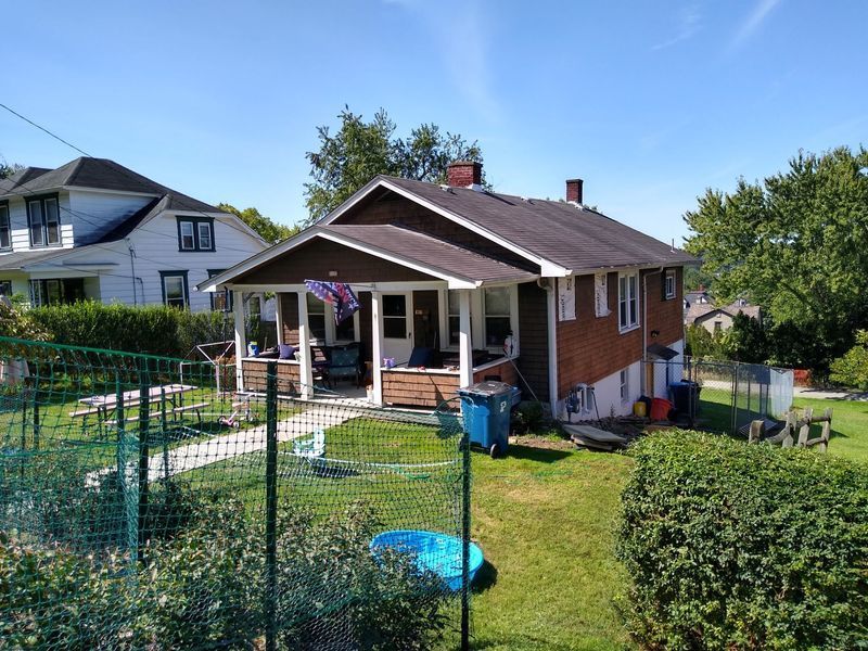 Small brick house with porch, green lawn, blue pool, and fence on a sunny day.