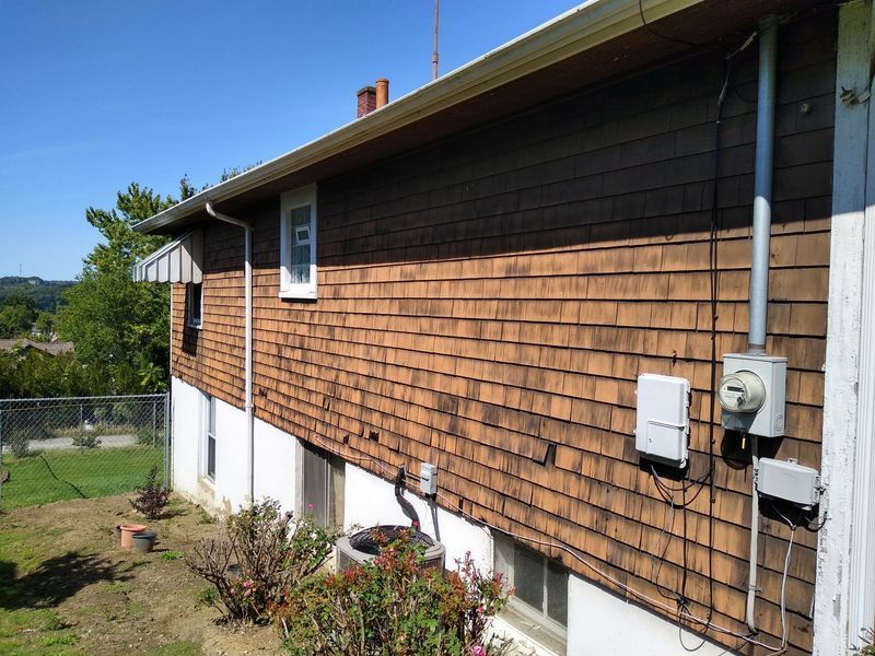 Brown shingle-sided house exterior with electrical panel, windows, and gutter against a blue sky.