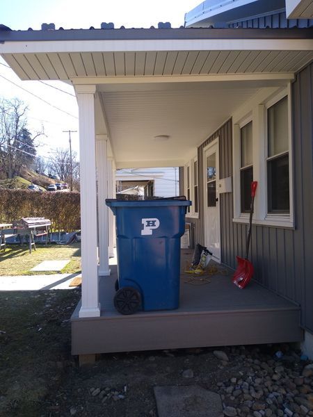 Blue trash bin on a gray porch under a white-trimmed roof. A red shovel leans against the building.