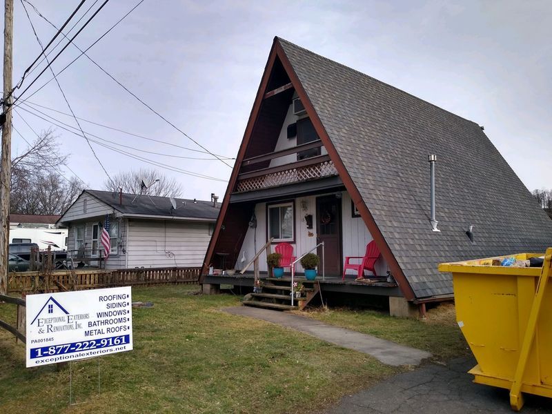 A-frame house with a brown roof and a small white house next to it, cloudy sky.